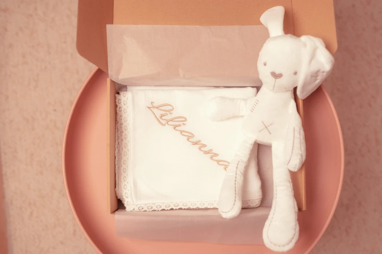 alt="Personalizowany mały zestaw prezentowy dla niemowlaka – muślinowa pieluszka z koronką oraz przytulanka królik z haftem imienia dziecka. Idealny prezent na chrzest i narodziny."